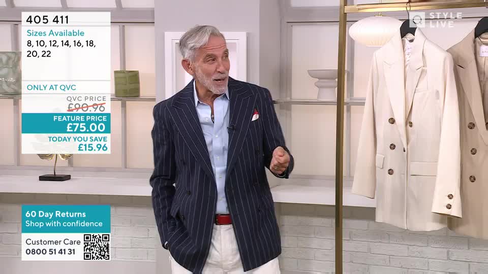 A man in a pinstriped blazer gestures with his right hand, talking enthusiastically. Behind him, a rack displays cream-colored double-breasted jackets, all part of a QVC Style Live presentation.
