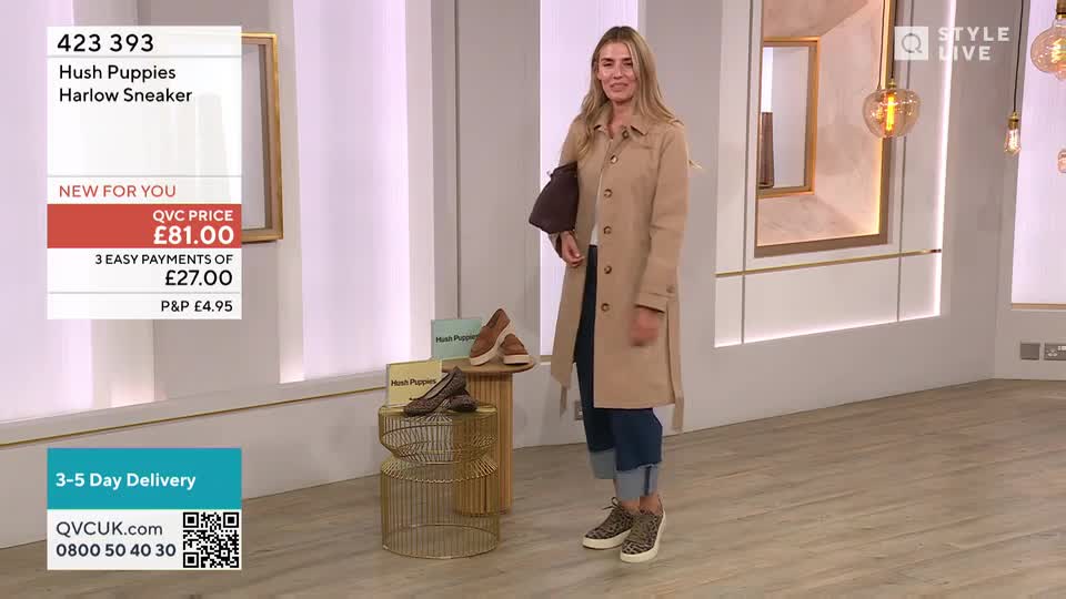 A woman in a tan coat and jeans stands in a studio, holding a bag and smiling at the camera.  She's wearing leopard-print sneakers, which are also displayed on a small table alongside other shoes.
