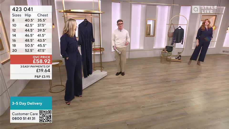 Two women and a man stand in a studio, with a navy jumpsuit on display. The woman on the left gestures towards the garment, while the man holds a script and looks towards the camera.
