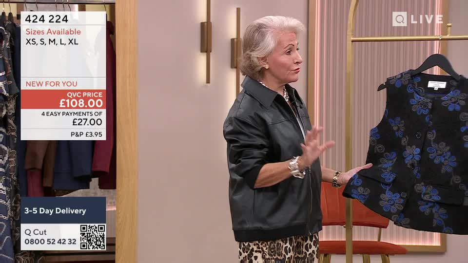 The presenter gestures towards a black vest with blue floral embroidery, held up on a hanger. She's wearing a black jacket and leopard-print trousers, standing in a well-lit studio.
