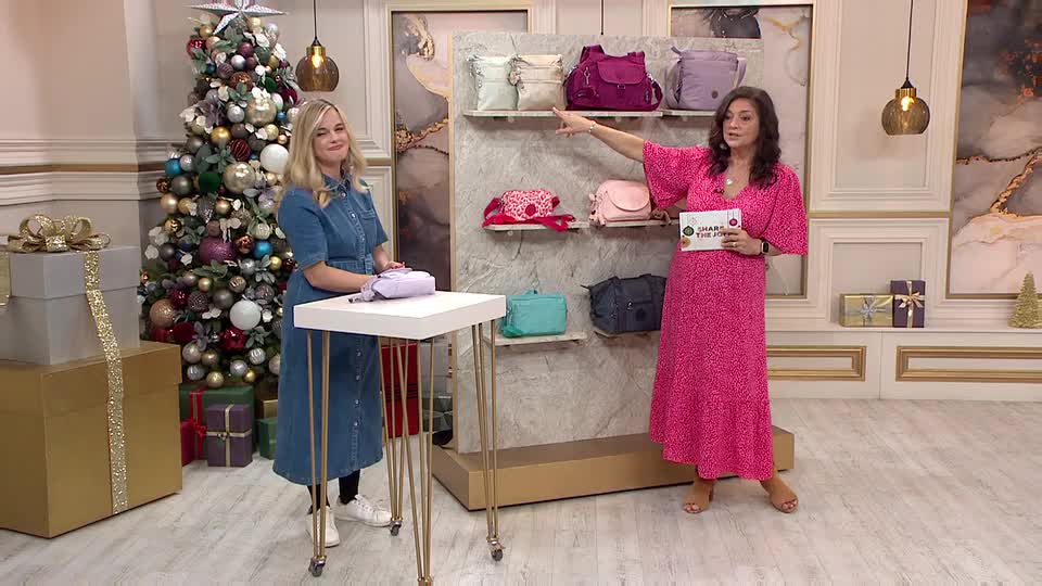A woman in a bright pink dress gestures towards a display of handbags, while her colleague in a denim dress stands nearby. The set has a Christmas tree and gift boxes, typical of a QVC-style presentation.
