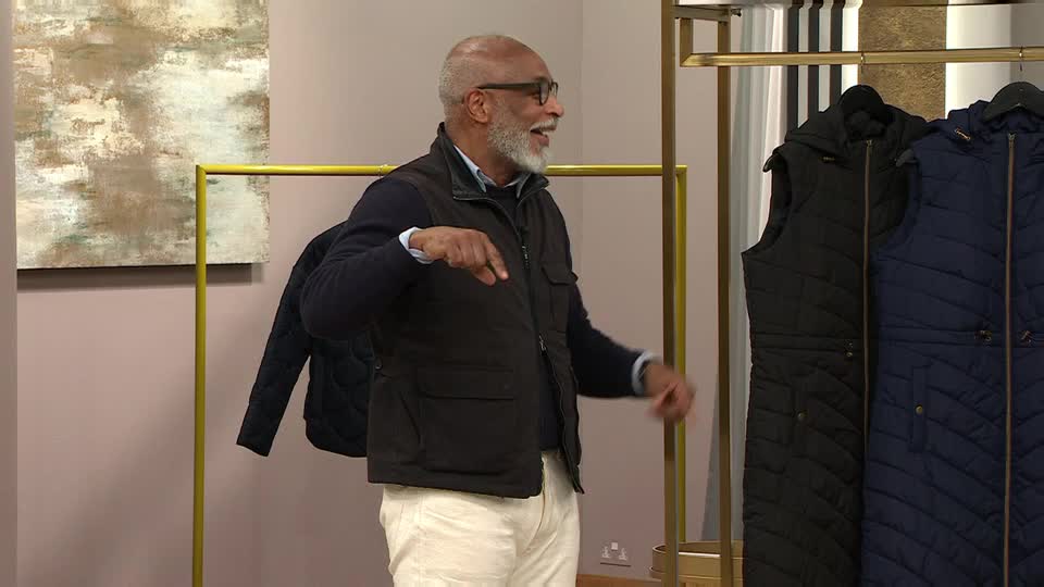 The man in the waistcoat gestures towards the rack of jackets, his expression animated. He's wearing a dark vest over a blue sweater, and the jackets on display are black and navy.
