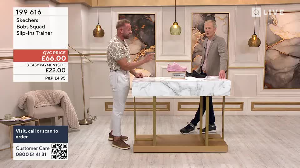 Two men stand at a marble-topped table, gesturing towards a pair of trainers. One man points at the shoes, while the other leans in, as if ready to offer more details on this QVC show.
