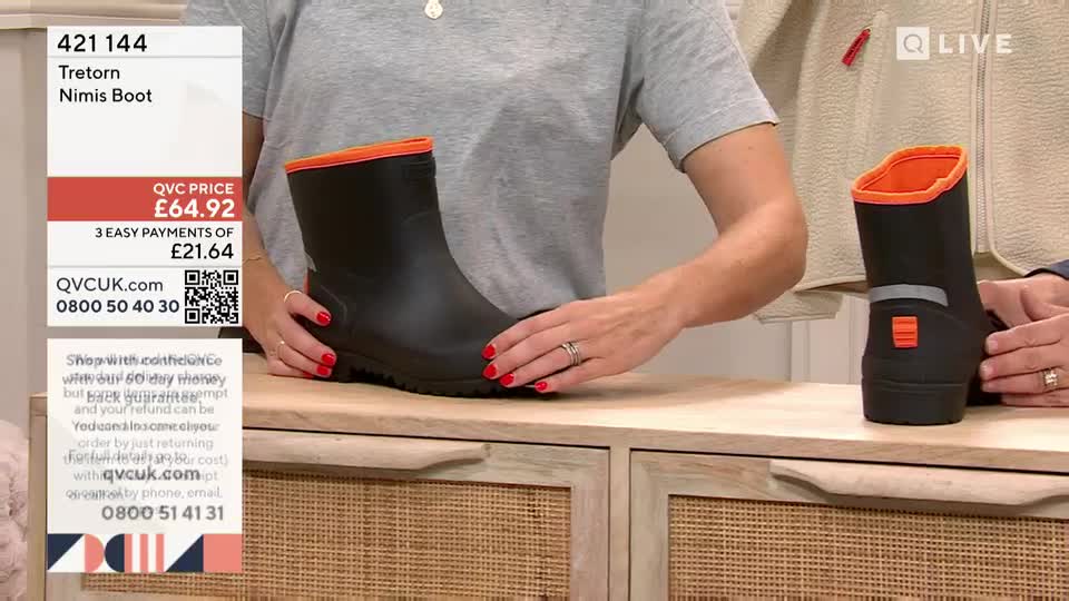 A woman in a grey t-shirt is holding a black wellington boot with an orange trim, showing it off to the camera. Another person is holding a matching boot, and the price is displayed on the screen.
