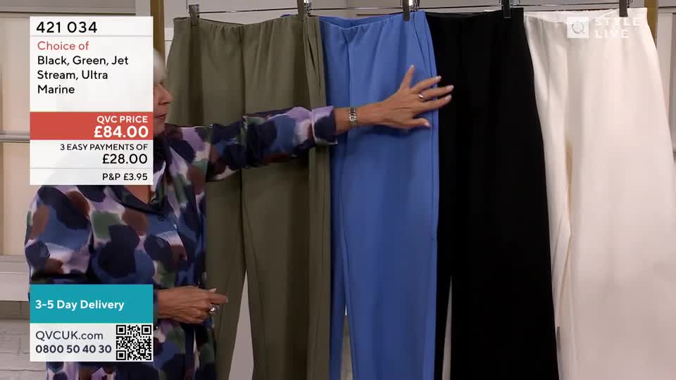 A presenter in a patterned top is touching the blue pair of trousers, showing the camera how the fabric feels. Behind her, four pairs of trousers in different colours hang from a rail, ready to be sold on QVC.
