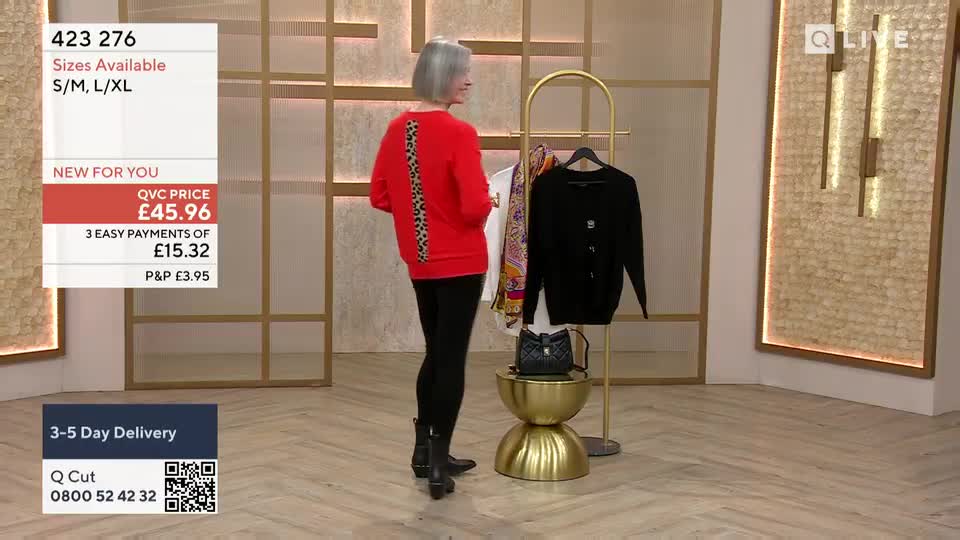 A woman in black leggings and boots stands facing away, modeling a red sweater with a leopard print stripe down the back. Beside her, a rack holds a black cardigan and a patterned scarf, all displayed in a QVC-style studio.
