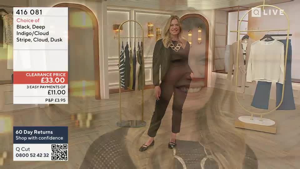 A presenter in a dark jumpsuit and a statement necklace smiles directly at the camera. Behind her, clothing hangs on gold racks, and the QVC UK price and product details are displayed on screen.
