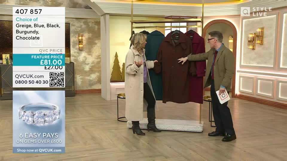 A woman in a long coat gestures, while a man in a blazer points at a row of coats hanging on a rack. The background suggests a QVC UK studio, with the price and colors listed on a screen.
