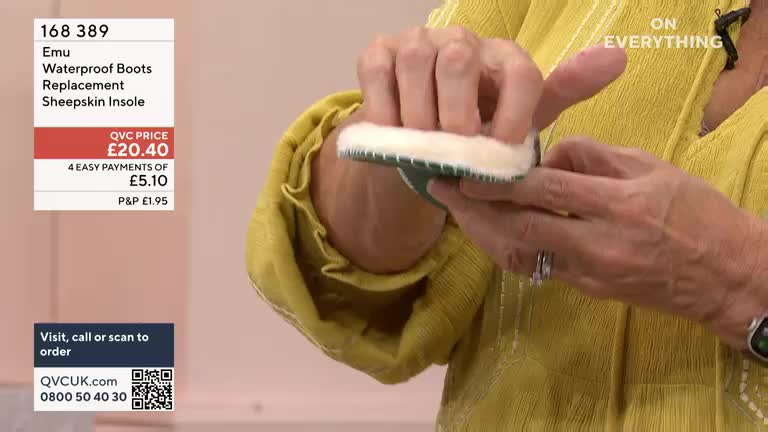 A woman holds a sheepskin insole, showing it to the camera on a QVC UK broadcast. She's pinching the fluffy white material between her fingers, highlighting its texture.
