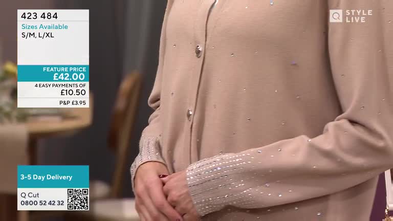 A woman stands with her arms crossed, showing off a beige cardigan on a QVC Style Live broadcast. Tiny sparkly embellishments dot the fabric, and the cuff is detailed with a row of them.
