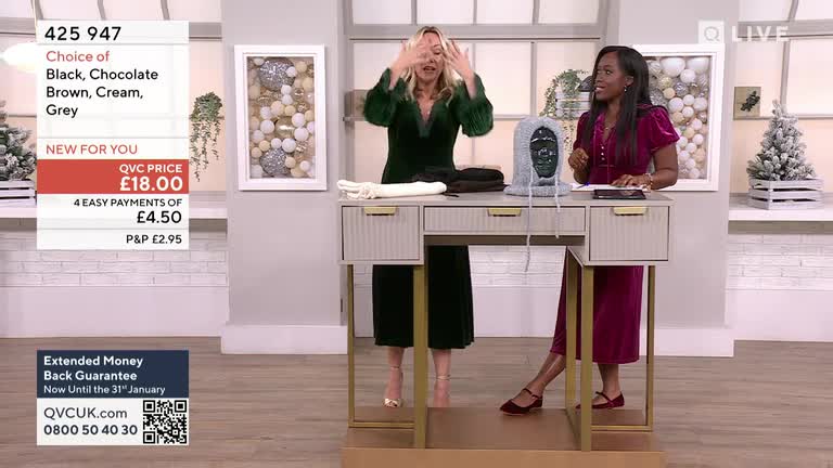 A woman in a green dress gestures with both hands as she stands behind a desk, while another woman in a magenta dress sits nearby. A knitted hood sits on the desk between them, and the QVC price is clearly displayed on the screen.
