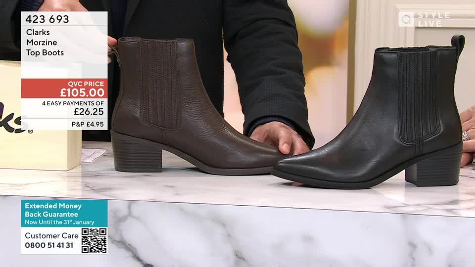 A brown leather boot sits next to a black one, both on a shiny surface. The presenter is holding the brown boot, showing it off for the QVC Style audience.
