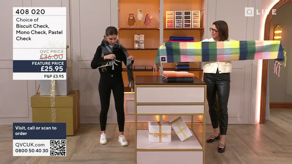 A presenter holds a large, colorful checked scarf, while another models a different scarf, tying it around her neck. The set is a well-lit QVC studio, with a display of scarves and gift boxes.
