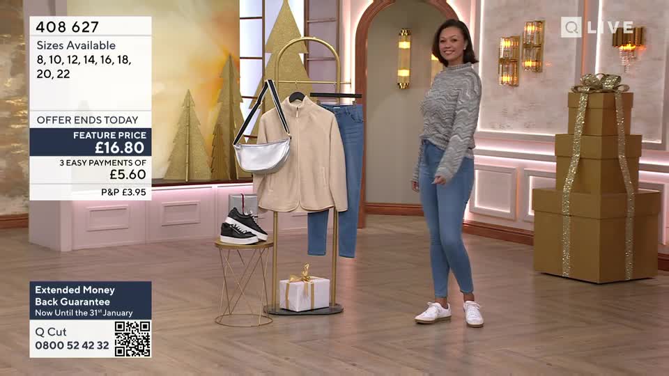 A woman in jeans and a sweater stands in a studio, smiling. Behind her, a rack displays a jacket, purse, and shoes, with a price prominently displayed on a screen.

