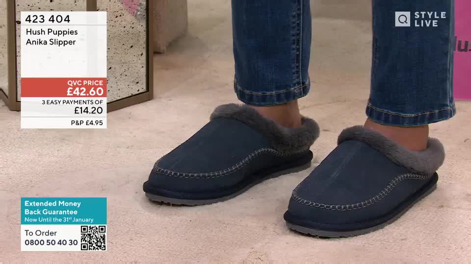 A person's feet are clad in dark blue Hush Puppies slippers, the fur trim visible around the ankles. The camera focuses on the footwear, displayed in a QVC-style presentation.
