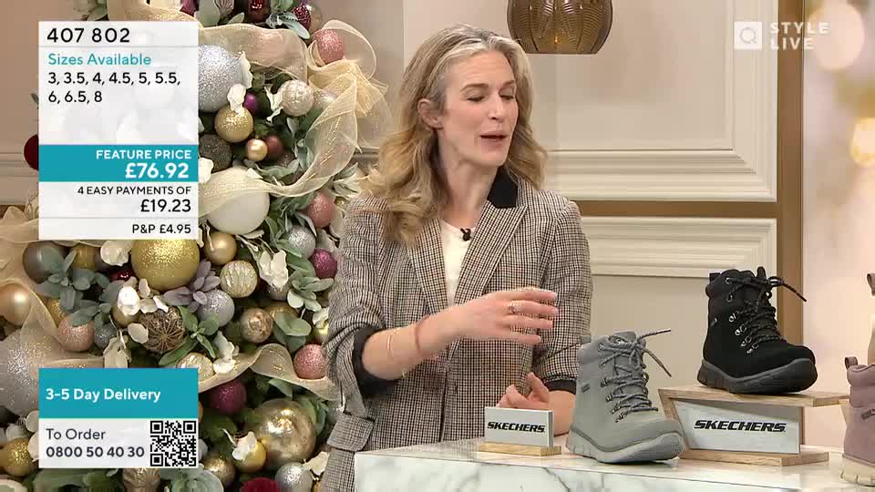A woman in a blazer gestures towards a selection of boots on display. Behind her, a Christmas tree glitters with ornaments, and a screen displays the price and ordering information.
A woman in a blazer gestures towards a selection of boots on display. Behind her, a Christmas tree glitters with ornaments, and a screen displays the price and ordering information.