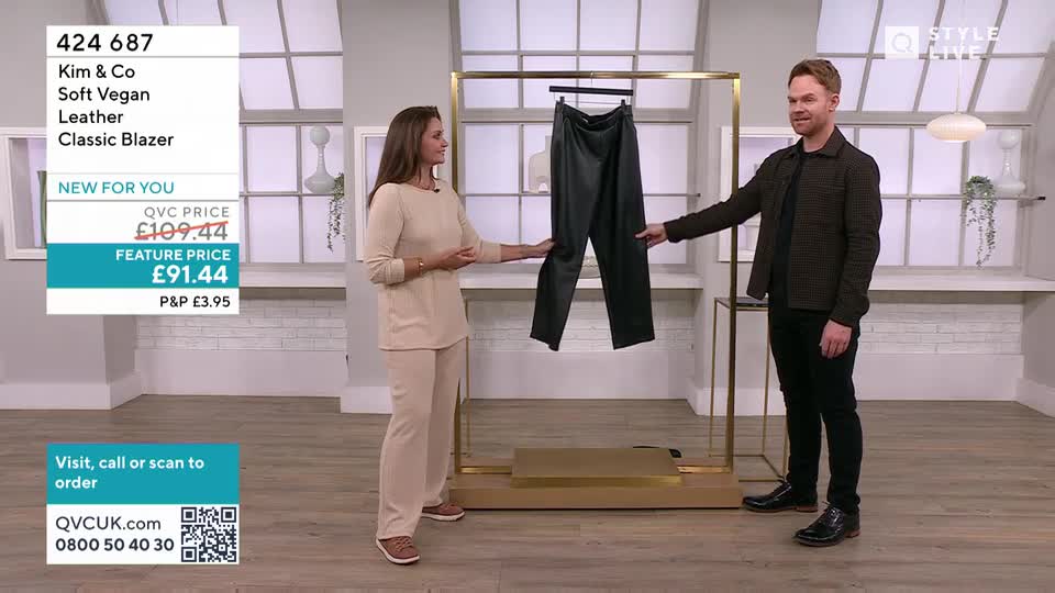 A woman in a beige outfit gestures towards a pair of black trousers hanging on a rack. A man beside her points to the same trousers, his hand near the waistband.