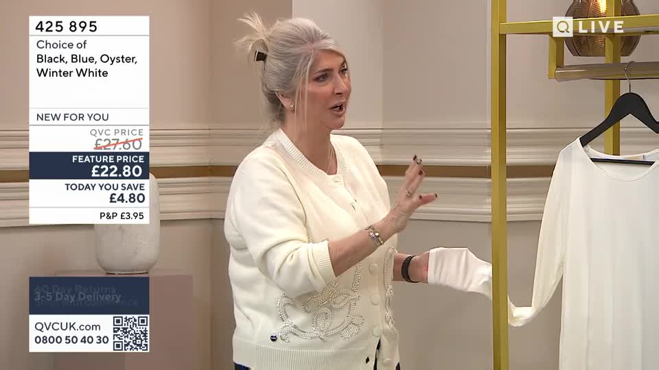 A woman in a cream cardigan gestures animatedly, holding a swatch of fabric. A white top hangs on a gold rack beside her.