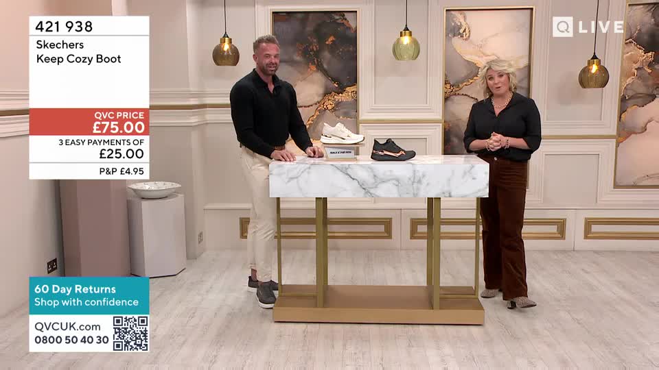 A man in a black shirt and cream trousers gestures towards a pair of white trainers on a marble-topped table. To his right, a woman in a black top and brown trousers stands with her hands clasped.