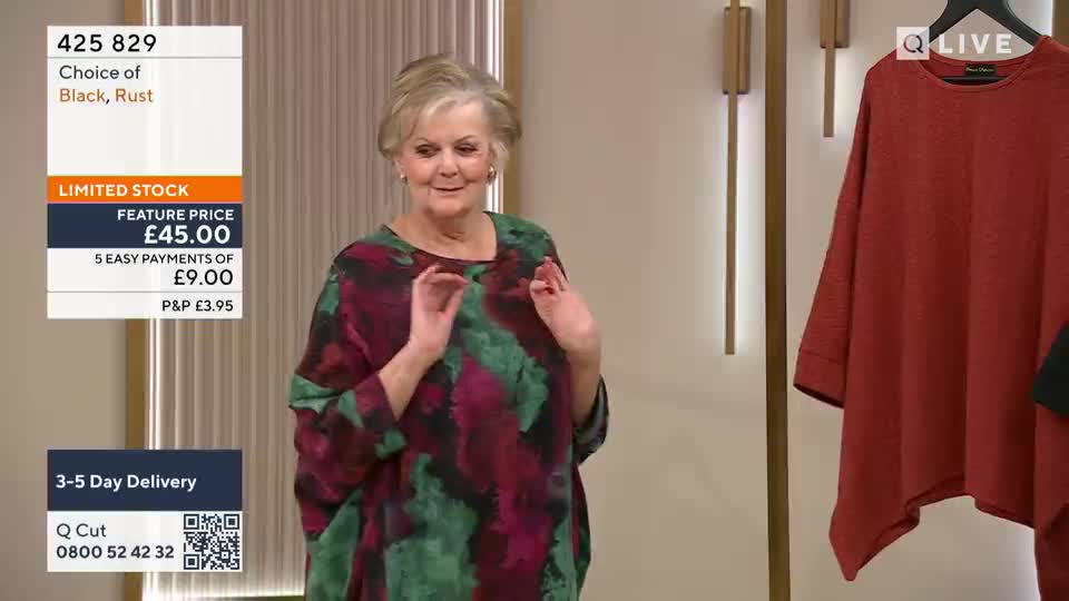 A woman in a colourful top gestures with her hands. Beside her, a rust-coloured garment hangs on a display.