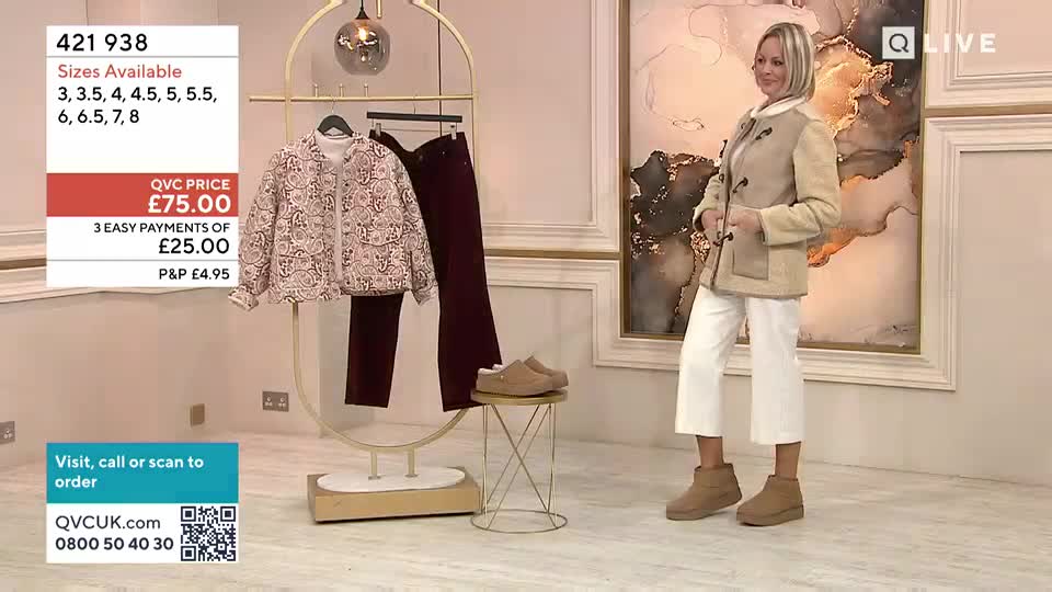 A woman stands in a studio, showcasing a light beige jacket over white trousers. On a nearby rack, a patterned jacket and dark trousers hang, with a pair of tan slip-on shoes resting on a small stool.