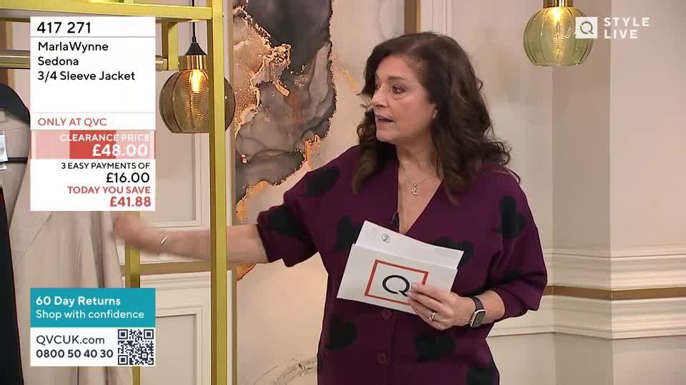 A woman in a burgundy cardigan with black polka dots gestures towards a display of clothing. She holds a card with a large "Q" on it, as seen on QVC.