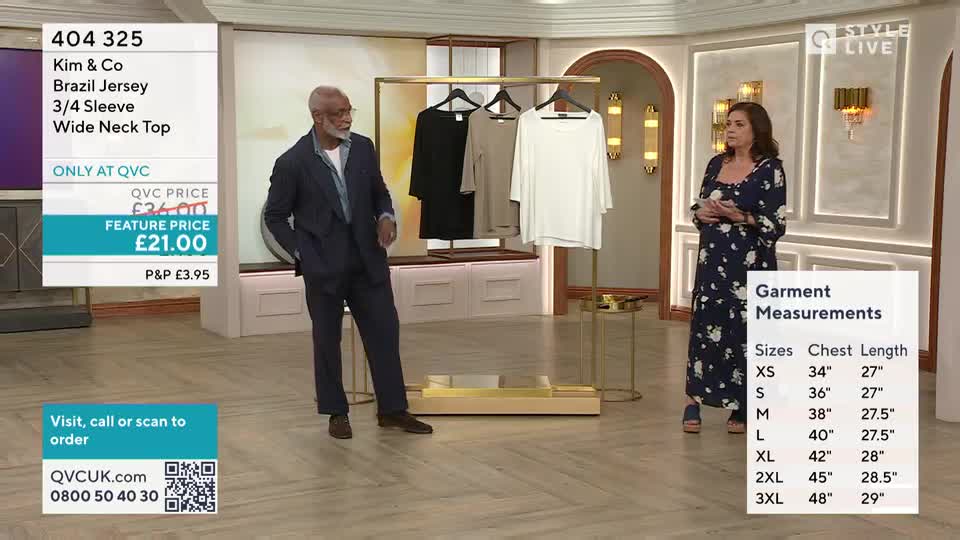 A man in a navy suit stands with his hands in his pockets, gesturing towards a rack of three tops. A woman in a floral dress stands to his right, looking at the camera.