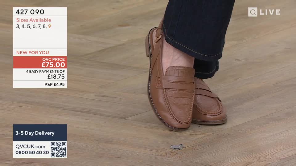 A person's feet, clad in brown leather backless loafers, are positioned on a light wood floor. The shoes are a classic penny loafer style, with a strap across the vamp.