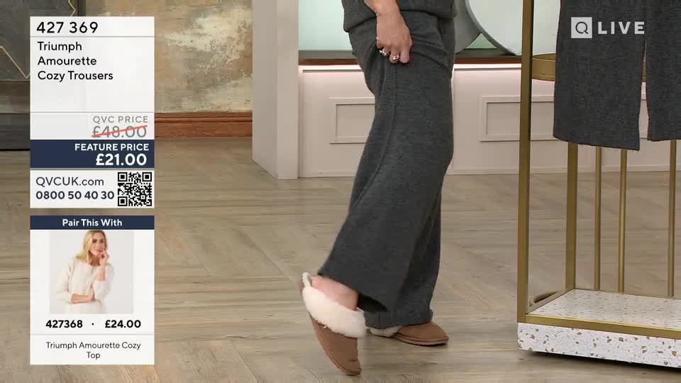 A woman demonstrates the fit of charcoal grey, wide-leg trousers by lifting one foot, revealing a fluffy slipper. The QVC logo and product details are displayed on a screen beside her.