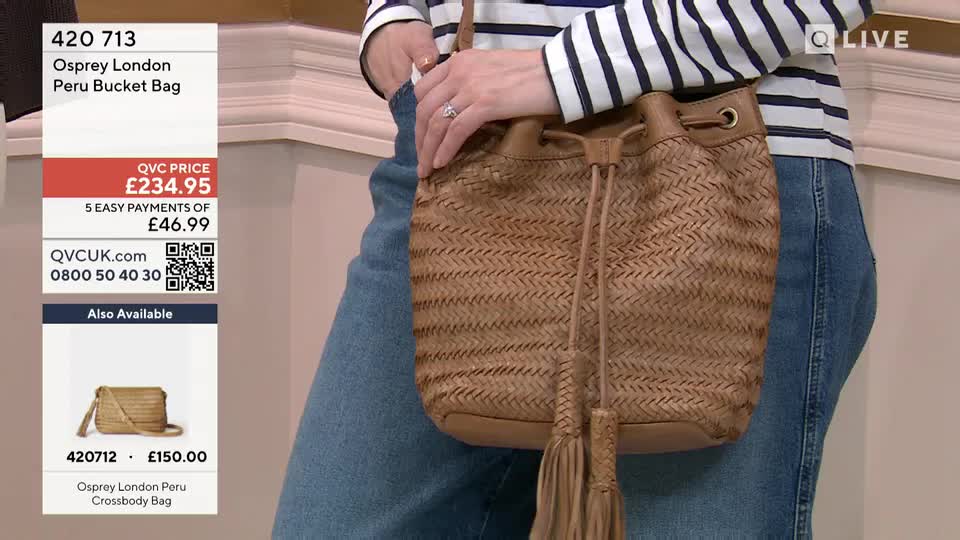 A woman in blue jeans holds up a tan, woven bucket bag with tassels. The bag is presented on a QVC broadcast, with prices and ordering information displayed on screen.