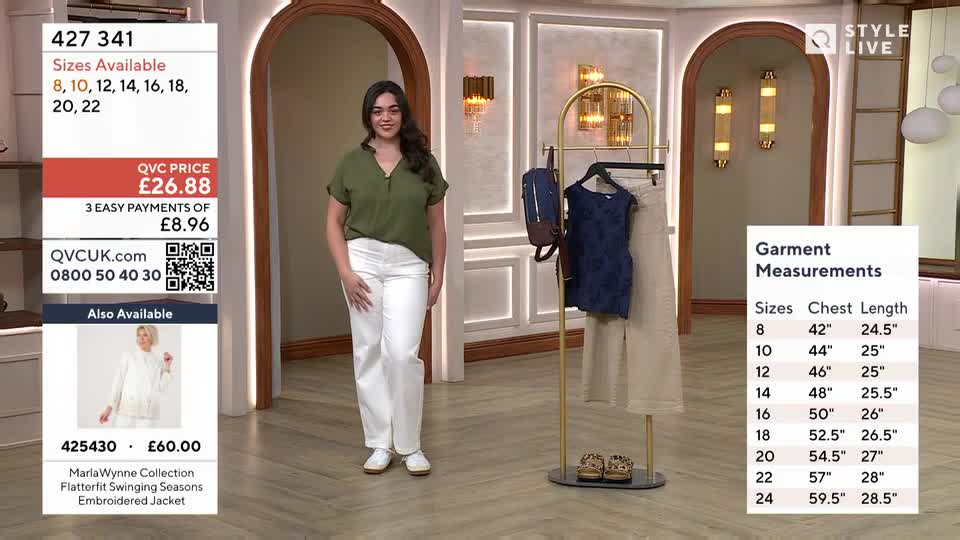 A woman stands in a studio, showcasing a green top and white trousers. On a nearby rack, a blue patterned dress and beige trousers hang, accompanied by a navy backpack.
