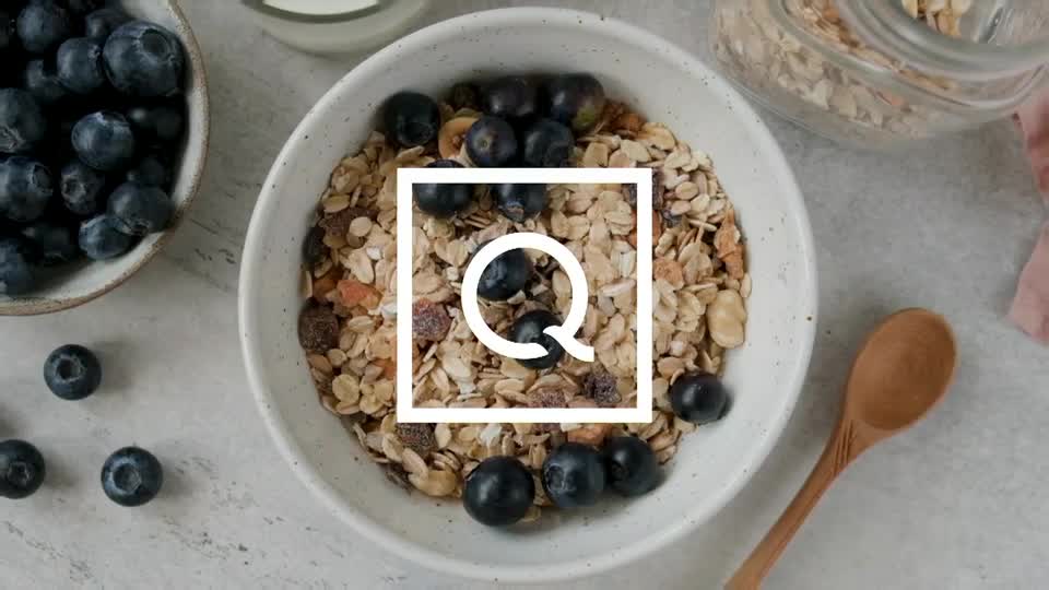 A bowl of muesli with blueberries sits on a light grey surface. Beside it, a small dish overflows with fresh blueberries, and a wooden spoon rests nearby.