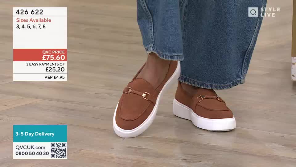 A pair of tan loafers with gold buckles sits on a wooden floor, their white soles catching the light. The wearer's wide-leg jeans are cuffed just above the shoes.