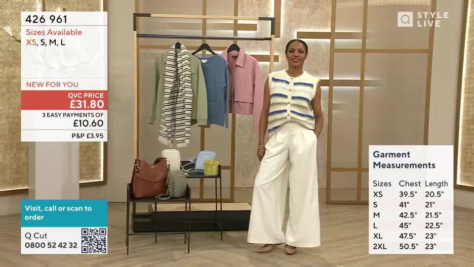 A woman in a striped sleeveless cardigan and wide-leg trousers stands on a television set. She's presenting clothing items displayed on a rack behind her.