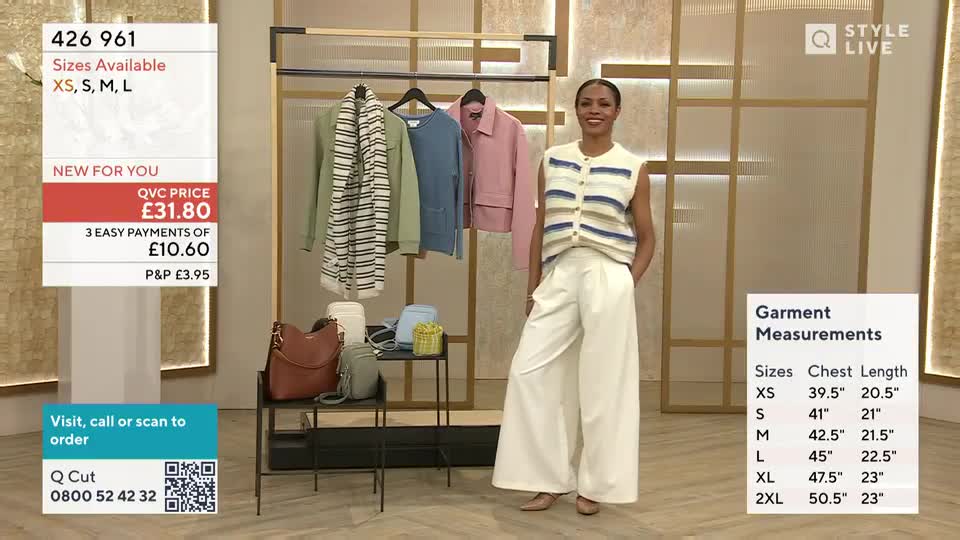 A woman stands on a stage, modelling a cream knit vest and wide-leg trousers. Behind her, a rack displays several garments, including a green jacket and a pink coat.
