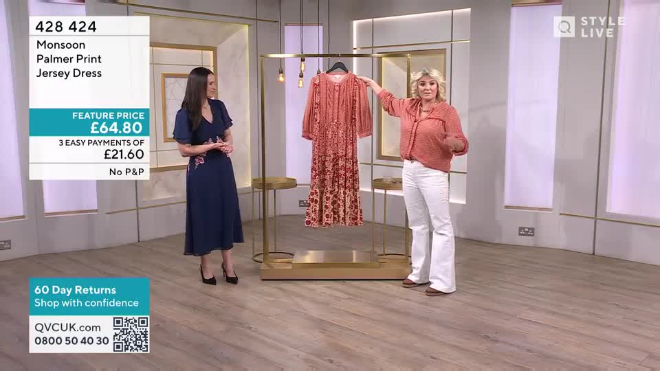 A woman in a coral blouse and white flared trousers gestures towards a coral-pink Monsoon dress hanging on a rack. Beside her, another woman in a navy dress stands with her hands clasped.