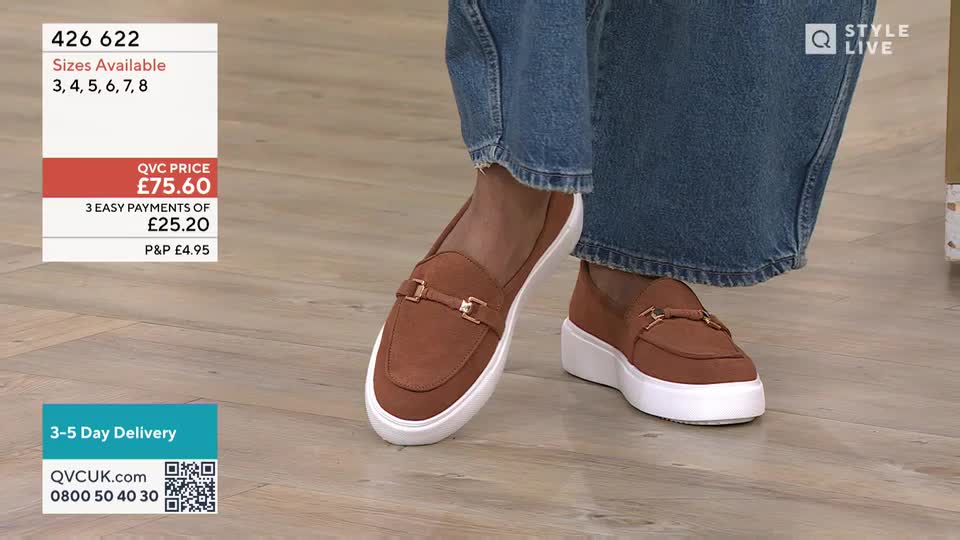 A pair of brown suede loafers with gold buckles sit on a wooden floor. The shoes have thick white soles and are paired with wide-leg denim jeans.