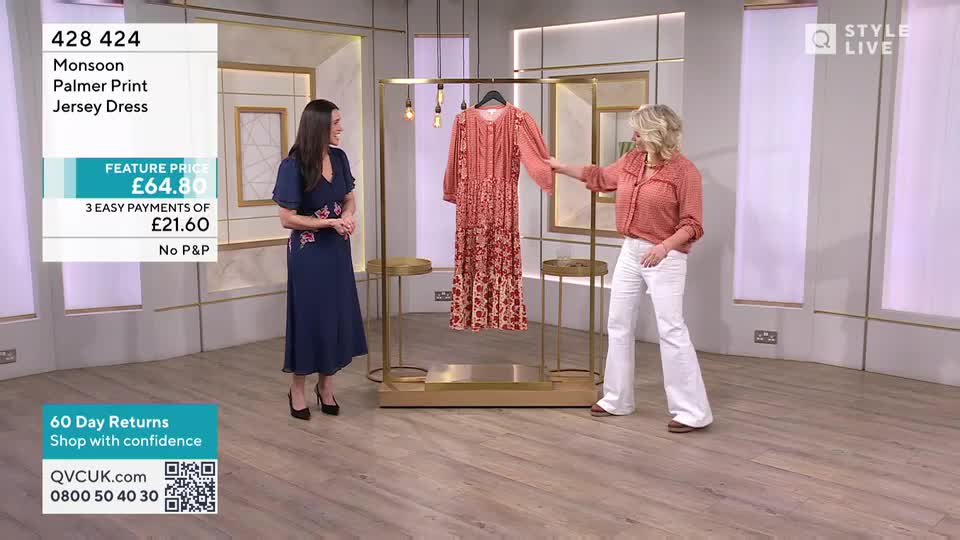 A woman in a coral blouse and white trousers gestures towards a patterned dress hanging on a display. Another woman in a navy dress stands nearby, smiling. A woman in a coral blouse and white trousers gestures towards a patterned dress hanging on a display. Another woman in a navy dress stands nearby, smiling.