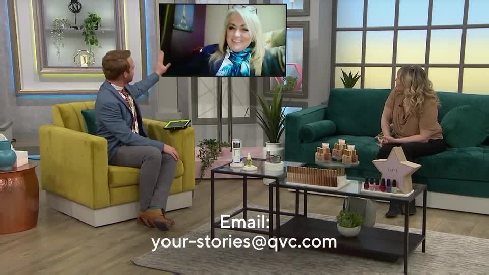 A man in a blue blazer gestures towards a screen showing a woman with blonde hair and a blue scarf. Beside him, a woman in a taupe top sits on a dark green sofa.