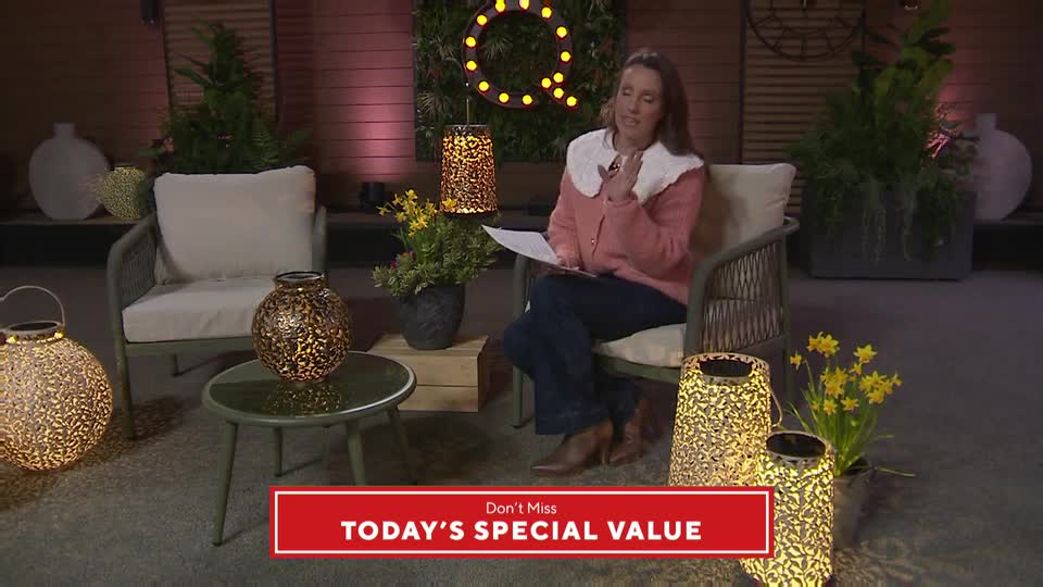 A woman in a pink cardigan sits on a chair, holding papers and speaking. Ornate, illuminated lanterns surround her, casting a warm glow on the scene.