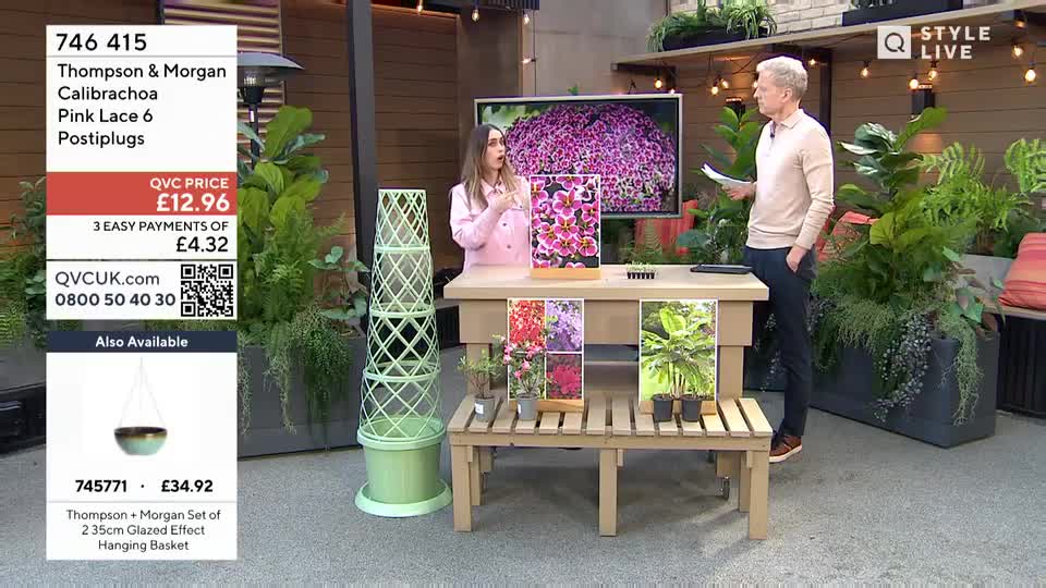 A woman in a pink jacket gestures as she speaks, while a man in a beige polo shirt stands nearby holding papers. They are presenting plants on a wooden table, with a large screen behind them displaying a vibrant purple flower.