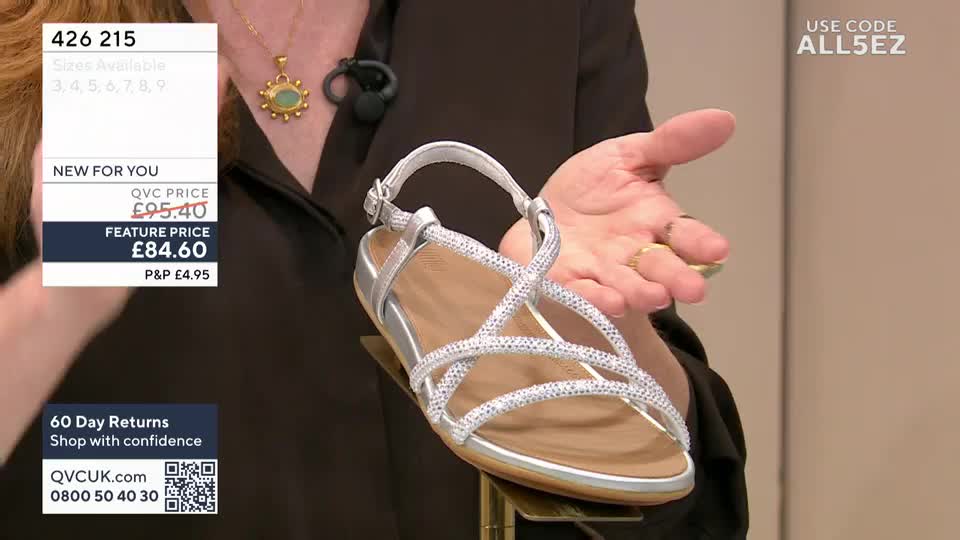 A silver sandal, its straps encrusted with tiny gems, is presented on a stand. The presenter's hand gestures towards the shoe, her rings catching the light.