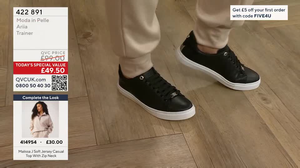 A pair of black trainers with white soles are being shown off, the feet moving slightly on a wooden floor. The QVC price and details are displayed on the screen in the United Kingdom.
