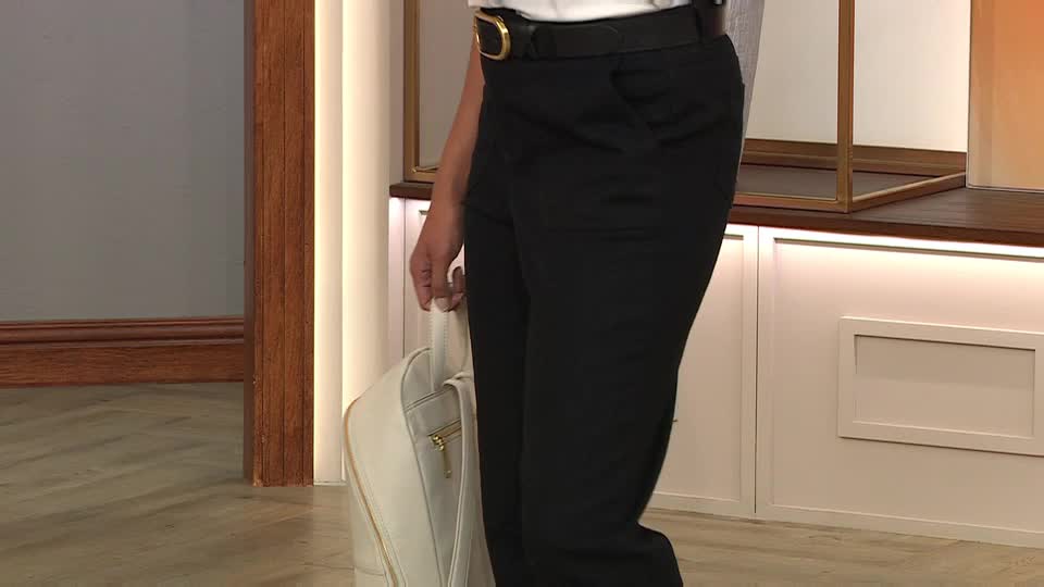 A person in black trousers and a white top is holding a white backpack. The gold buckle on their belt catches the light in the studio.
