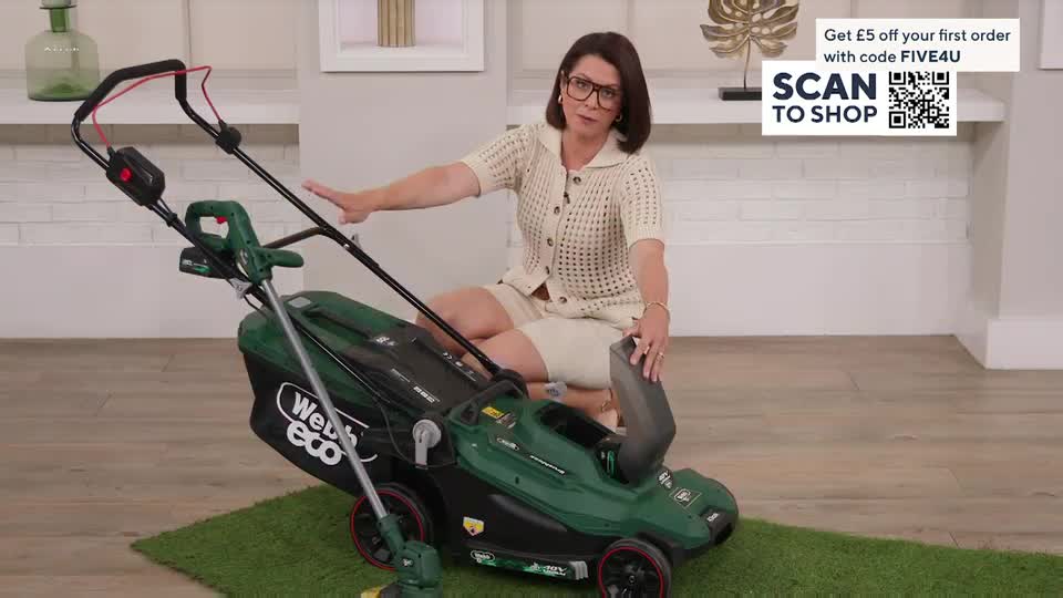 The woman, seated on the floor, gestures towards a green lawnmower, her hand resting on its handle. A smaller grass trimmer stands to the side, while a QVC graphic in the background offers a discount.
