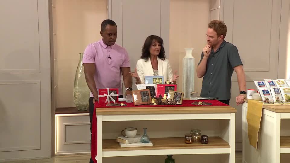 A woman in a white blazer gestures towards a table laden with gift boxes and framed photos. The man in the pink shirt stands to her left, while the other man, in a gray shirt, strokes his chin, seemingly pondering.
