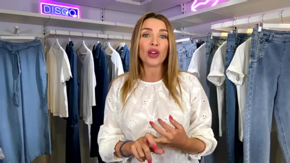 A woman in a white eyelet blouse gestures with her hands, speaking animatedly in front of racks of denim and white tops. Above, a neon sign spells out "DISCO" in a bright pink glow.
