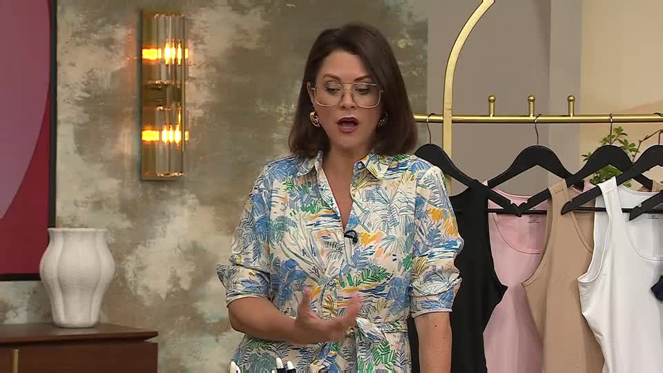 The woman in the floral shirt gestures with her hands as she speaks, likely on a QVC set in the UK. Behind her, a rack displays various tank tops, ready for presentation.
