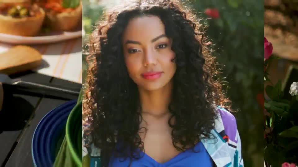 A woman with dark, curly hair is framed in the center, wearing a blue top and a colorful jacket. The scene looks like a segment on QVC, with food and flowers visible on either side.
