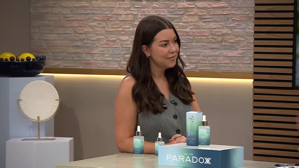A woman sits at a table, looking to her right with a slight smile. Beside her, several bottles and boxes of "Paradox" skincare products are neatly arranged.
